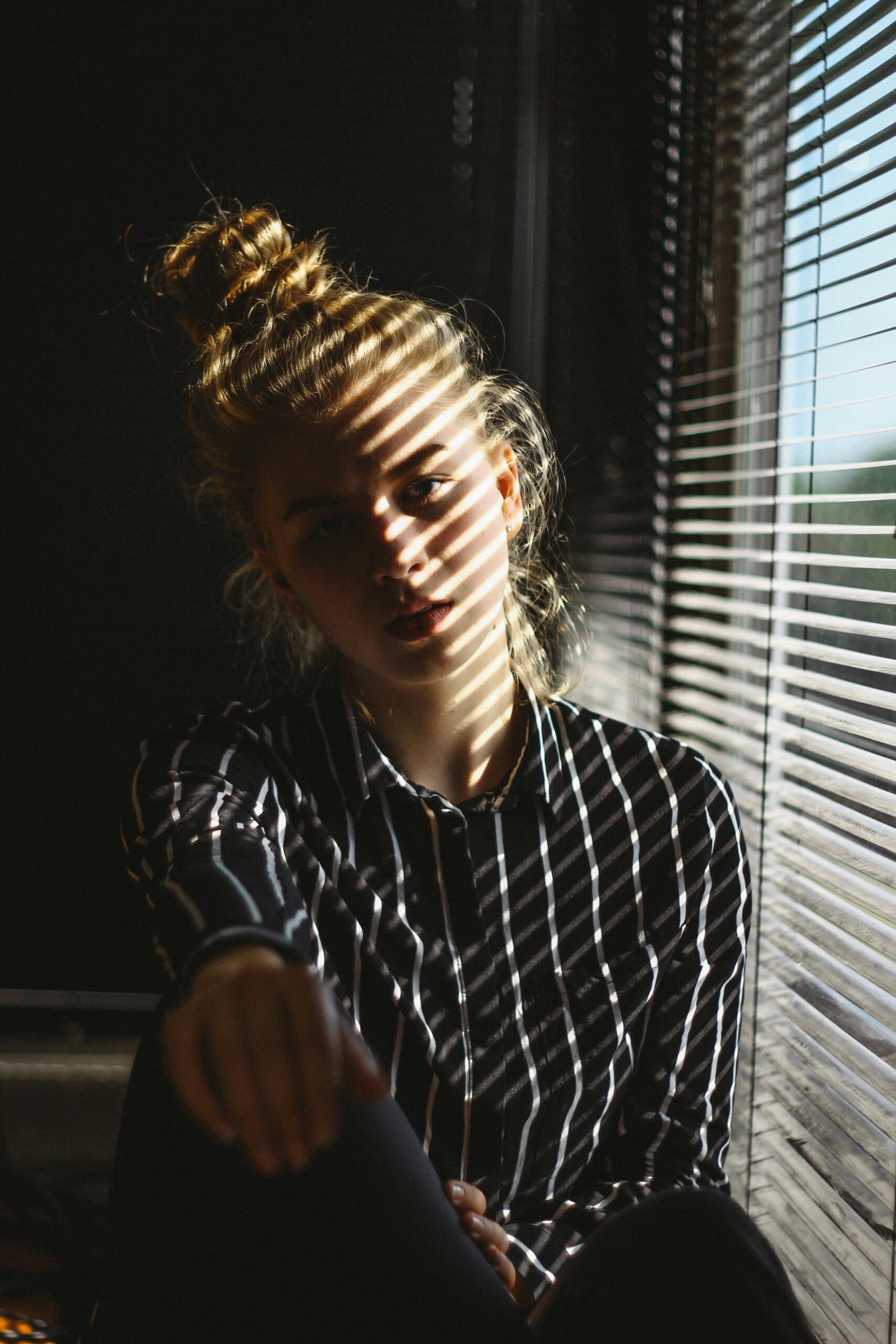 A contemplative woman sits by window blinds casting shadows inside.